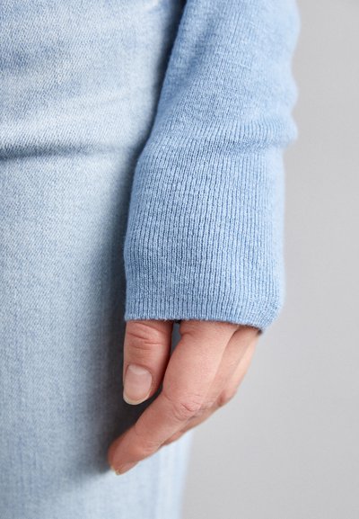 Käsi rentona sivulla, päällä vaaleansininen neule, jossa on peukalonrei'itys, sekä vaaleansiniset farkut.