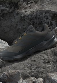 Chaussure de trail running gris foncé avec semelle texturée reposant sur un terrain accidenté et rocheux, mettant en avant ses caractéristiques de design extérieur et robuste.