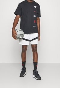 Camiseta negra de Nike con el logo de los Chicago Bulls; pantalones cortos de malla blancos con acentos negros; zapatillas deportivas negras; sosteniendo un balón de baloncesto gris.