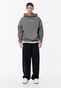 Młody mężczyzna stoi w szarej bluzie z kapturem z napisem „Kochaj siebie. Nie mnie”, w luźnych czarnych spodniach i białych sneakersach na białym tle.