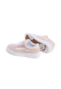 Une paire de sneakers en daim rose clair avec des accents blancs, des semelles blanches et un profil de semelle de couleur gomme, positionnée avec une chaussure sur le côté.