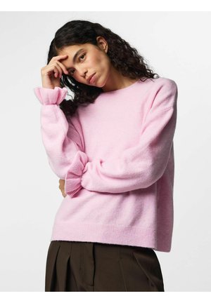 Femme aux cheveux bouclés foncés portant un pull rose avec des poignets à volants et un pantalon marron foncé, posant avec la main sur le front sur fond blanc.