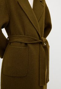 Olivegrön ullkappa med sjalkrage, omslagsdesign, knytbälte och två framfickor med en mjuk, konstig yta.