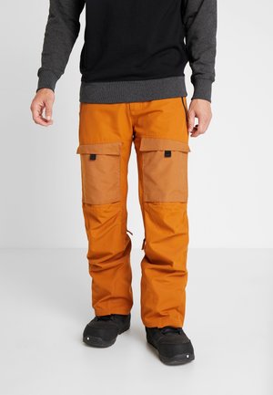 Mand iført lyse orange cargobukser med store forlommer og sorte sko, stående mod en ensartet lys baggrund.