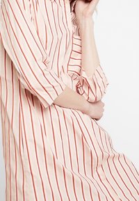 Vestido rosa claro con finas rayas rojas, presenta un cuello y mangas tres cuartos. Tejido suave, corte holgado, diseño hasta la rodilla.