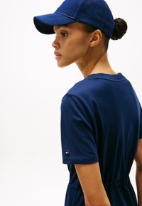 Robe en coton bleu marine à manches courtes et taille avec cordon de serrage, avec un petit logo sur la manche. Casquette à visière incurvée dans la même couleur.