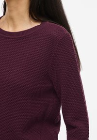 Burgunderfarget strikkegenser med teksturert mønster, rund hals og lange ermer. Stoffet ser mykt og noe elastisk ut.