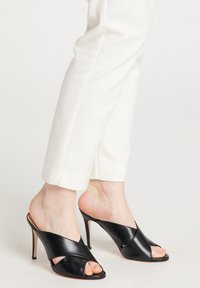 Zwarte hoge muiltjes met gekruiste banden, open te ontwerp, gladde lederen textuur en een slanke stilettohak. Gecombineerd met witte broek.