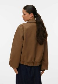 Veste en polaire marron à coupe décontractée, manches longues, et ourlet froncé. Dotée d'un col et d'une texture lisse. Portée par-dessus un jean denim foncé.