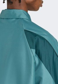 Türkise Jacke mit einer glatten, satinartigen Textur, ausgestattet mit einem strukturierten Kragen und einem kontrastierenden, dunkleren türkisen Einsatz entlang der Schulter.