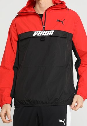 PUMA windbreaker jacket featuring a red upper section and black lower section, with a prominent white logo, zip neck, and elastic cuffs.