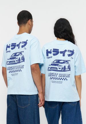 Dwie osoby noszące jasnoniebieskie koszulki z niebieską grafiką samochodu i napisem „Tokyo Night Drive” na plecach.