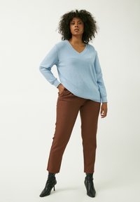 Helles blaues V-Ausschnitt-Pullover mit gerippten Bündchen, kombiniert mit braunen schmal geschnittenen Hosen mit seitlichen Schlitzen und schwarzen ankle boots mit Absatz.