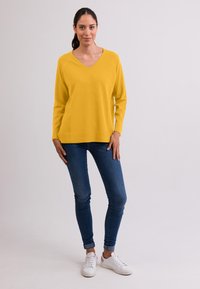 Žlutý svetr s výstřihem do V, dlouhými rukávy, měkkou texturou, ve spojení s modrými skinny džíny a bílými teniskami. Jednoduchý, ležérní design s pohodlným střihem.