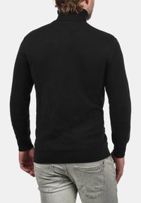 Schwarzer Strickpullover mit hohem Kragen, strukturiertem Muster und gerippten Bündchen und Saum. Getragen mit hellen Jeans, die Rückansicht wird gezeigt.