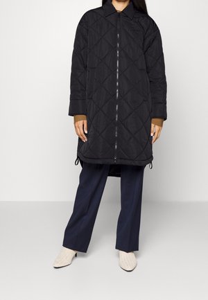Personne portant un long manteau matelassé noir, un pantalon large bleu marine, un haut à manches longues marron, et des bottines blanches texturées à talons.