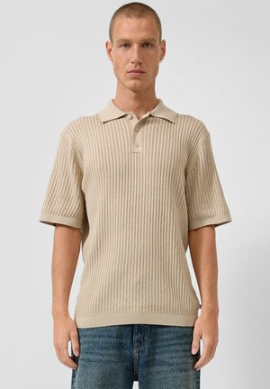 Junger Mann mit kurzen Haaren, der ein beiges, geripptes Polohemd mit kurzen Ärmeln und blaue Jeans trägt, vor einem schlichten weißen Hintergrund stehend.