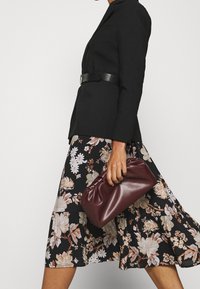 Schwarze Blazer mit einem Ledergürtel, schwarzer Rock mit Blumenmuster in Creme und Braun, und eine bordeauxfarbene Clutch, die in der Hand gehalten wird.