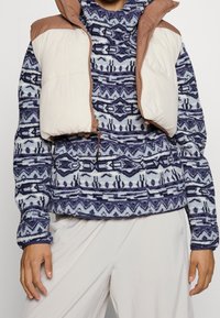 Gilet imbottito con sezioni beige e marrone, indossato sopra un top in pile blu a motivo geometrico. Texture liscia, forma aderente.