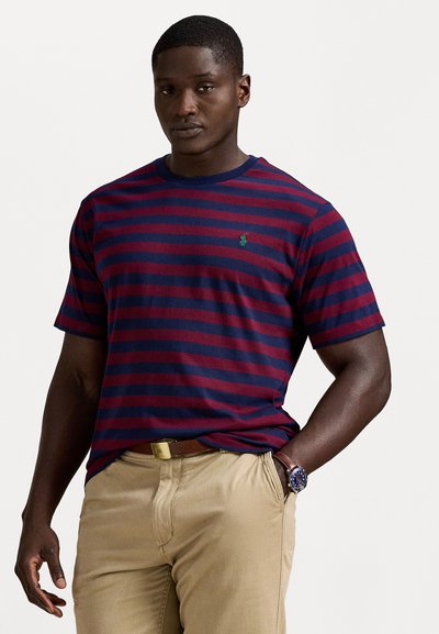 Gestreiftes T-Shirt in Marineblau und Burgunderrot aus Baumwolle, mit einem Rundhalsausschnitt und einem kleinen grünen Logo-Detail auf der linken Brust. Kombiniert mit beigen Hosen.