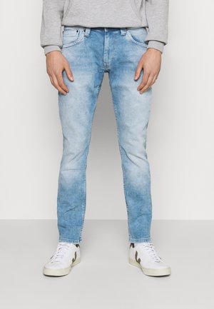 Man draagt lichtblauwe slim-fit jeans, grijze trui en witte sneakers met zwarte accenten, staand tegen een effen achtergrond.
