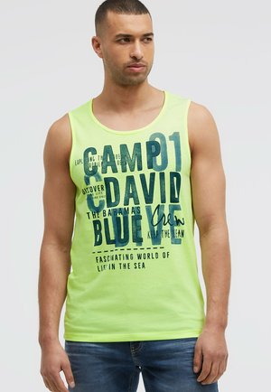 Mann trägt ein limettengrünes ärmelloses Shirt mit dunkelblauem Schriftzug einschließlich "Camp David" und Jeans, steht vor einem schlichten hellen Hintergrund.