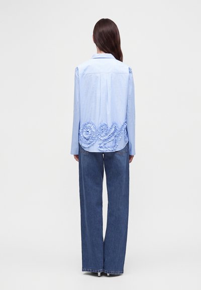Femme aux longs cheveux foncés portant une chemise rayée bleu clair avec une broderie florale volantée dans le dos et un jean bleu à jambes larges.