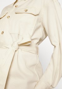 Gros plan d'une veste beige à manches longues avec poches à rabat devant, boutons en écaille de tortue et ceinture en tissu nouée en nœud à la taille.