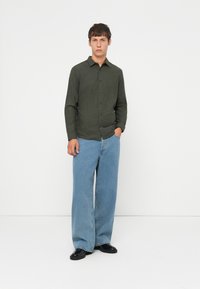 Camicia button-up verde scuro con colletto, abbinata a jeans a gamba larga blu chiaro e scarpe nere. Design semplice, vestibilità casual e tessuto strutturato.