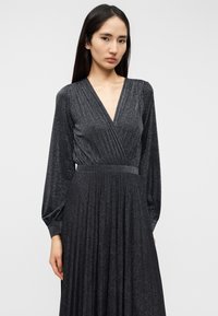 Μακρύ μαύρο φόρεμα με λαιμόκοψη σε σχήμα V, φτιαγμένο από υλικό με υφή και λαμπερό φινίρισμα. Διαθέτει μακριά, φουσκωτά μανίκια και εφαρμοστή μέση, που ρέει σε μια πλισέ φούστα.