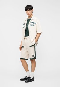 Νεαρός άνδρας που φοράει κρέμ κοντομάνικο πουκάμισο και σορτς με πράσινη διακόσμηση, πράσινη μπλούζα, άσπρες κάλτσες και μαύρα μοκασίνια, μπροστά σε λευκό φόντο.