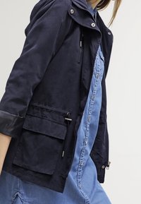 Granatowa lekka kurtka z kapturem, zapinana na zamek błyskawiczny, z dwoma przednimi kieszeniami. Charakteryzuje się gładką fakturą i kontrastującymi czarnymi mankietami na rękawach.