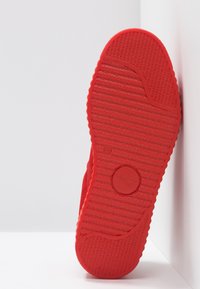 Semelle de chaussure rouge avec des motifs antidérapants texturés et une zone circulaire pour le logo, photographiée sur fond blanc.