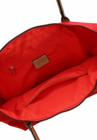 Red canvas tote bag with brown leather accents, zip closure, interior pocket, and smooth texture. Features durable stitching and a spacious interior.