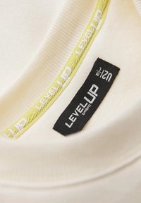 Tessuto color crema con una texture a righe presenta un accento verde e un'etichetta nera con la scritta "LEVEL UP" e la taglia "S/M."