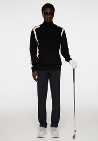 Svart zip-up tröja med vita sidränder, kombinerad med mörka byxor och grå sneakers. Accessoarer med solglasögon och en golfklubba.