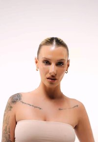Une femme aux cheveux raides et blonds clairs porte un haut beige sans manches. Elle a des boucles d'oreilles en forme de créole et des tatouages visibles sur les bras. Sa peau paraît lisse et lumineuse.