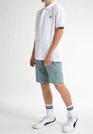 Niño de pie vistiendo una camiseta polo blanca, pantalones cortos de color teal, calcetas blancas con rayas negras y zapatillas Puma blancas sobre un fondo blanco.