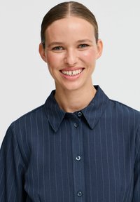 Camicia a righe blu navy con colletto e chiusura a bottoni, caratterizzata da una texture liscia e un tessuto leggero.