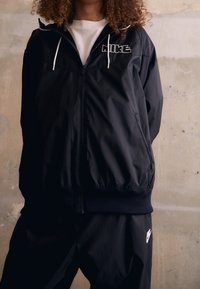 Svart Nike vindjacka med dragkedja fram, snörning i huvan och logotyp. Mjuk, lättviktsmaterial med elastiska ärmslut och nederkant.