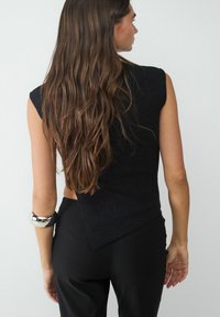 Schwarzes, geripptes, ärmelloses Top mit asymmetrischem Saum, getragen mit schwarzen Hosen und einem silbernen Armschmuck, der einen strukturierten Stoff präsentiert.