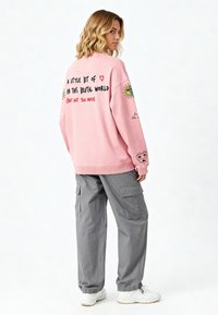 Svijetloružičasti dukserica s grafičkim tekstom na leđima i ilustracijama, uparen sivoj cargo hlačama, prikazuje opušteni kroj i manžete.