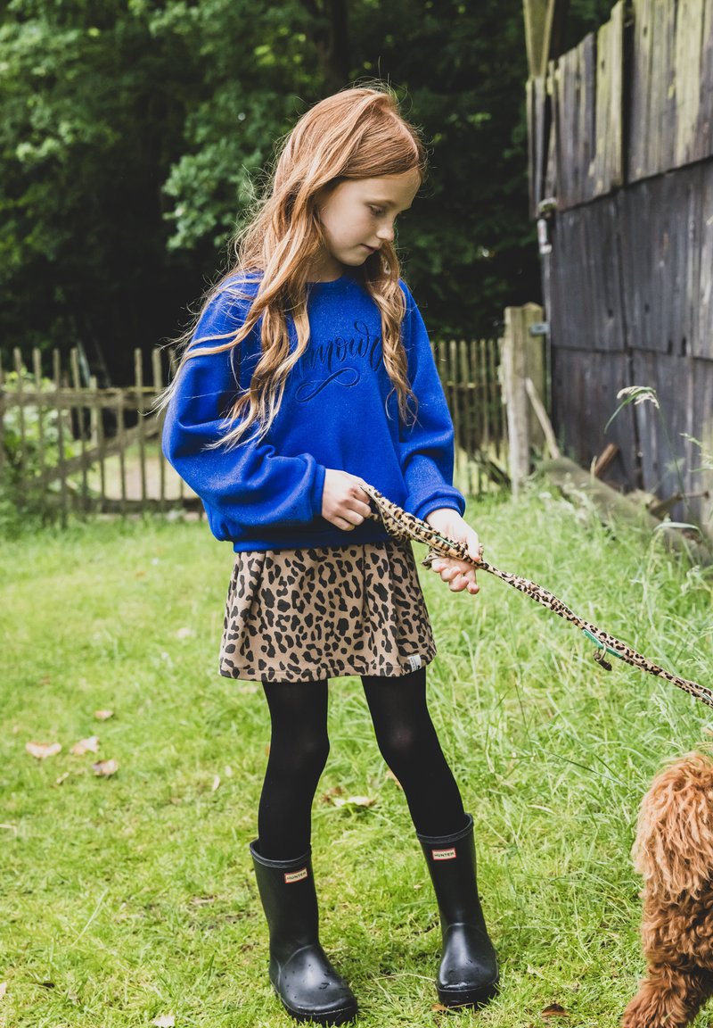Blauw sweatshirt met cursieve tekst, leopardprint rok, zwarte panty's en zwarte regenlaarzen. Het kind houdt een gepatternde hondenriem vast.