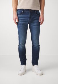 Sötétkék farmernadrág slim fit szabásban, finom fakulással, öt zsebes kialakítással és hagyományos gombos zipzárral. Fehér sportcipő tartozik hozzá.