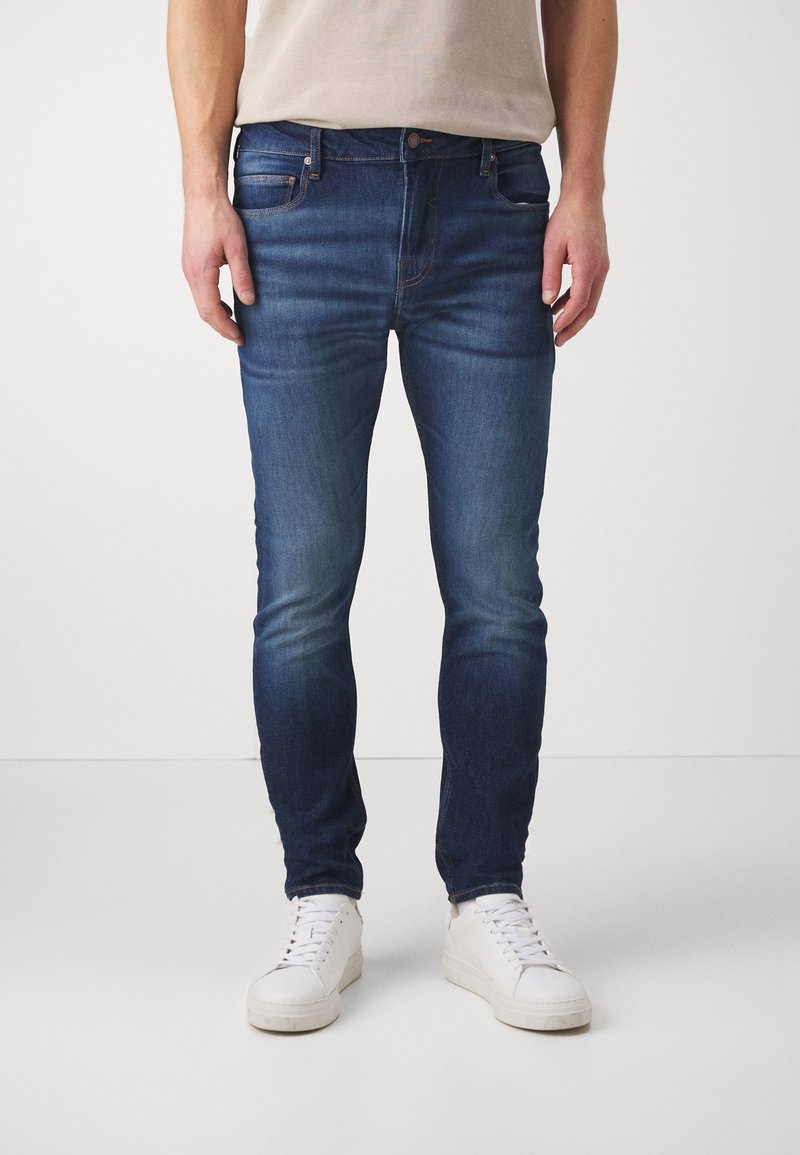 Sötétkék farmernadrág slim fit szabásban, finom fakulással, öt zsebes kialakítással és hagyományos gombos zipzárral. Fehér sportcipő tartozik hozzá.