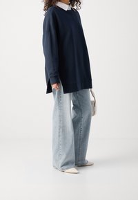 Sudadera de gran tamaño en color marino, de mangas largas, con aberturas laterales; jeans de corte ancho en azul claro; zapatos blancos de punta; bolso minimalista.