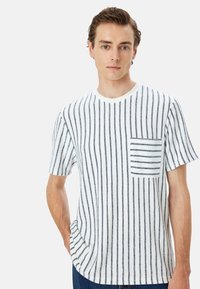 Camiseta blanca de manga corta con rayas verticales en navy y un bolsillo pequeño con rayas horizontales. Corte casual y escote redondo.