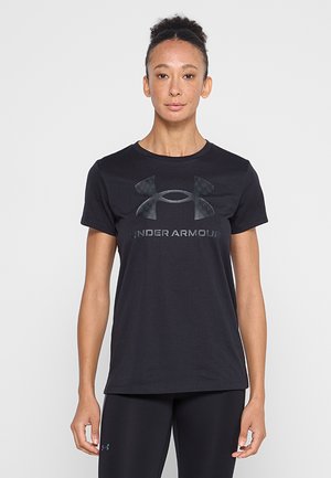 Młoda kobieta o naturalnie kręconych włosach nosi czarną koszulkę Under Armour i czarne legginsy, stoi na jasnym jednolitym tle.
