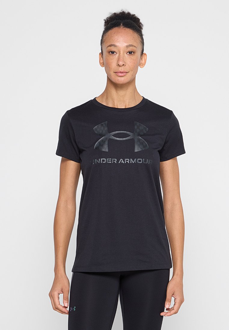 Jeune femme aux cheveux naturellement bouclés porte un t-shirt noir Under Armour et des leggings noirs, se tenant devant un fond clair uni.