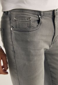 Jeans en denim gris avec une coupe slim, un design classique à cinq poches et des accessoires en ton argenté. La texture est lisse avec une légère décoloration.